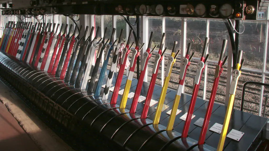 A long row of red, yellow, blue and black levers in a sunlit railway signal box, numbered plates on the nearest, the tracks outside its windows