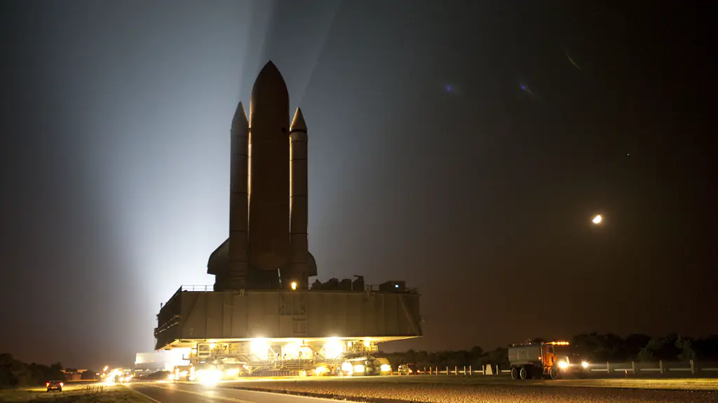 A space shuttle on its crawler moves along a lit road at night, backlit by floodlights, the moon at right