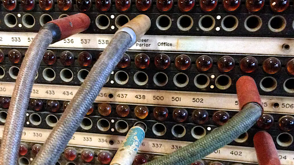 Fabric-covered cords with red, cream and blue plugs patched into the numbered jacks of an old hotel telephone switchboard, between rows of red and amber lamps